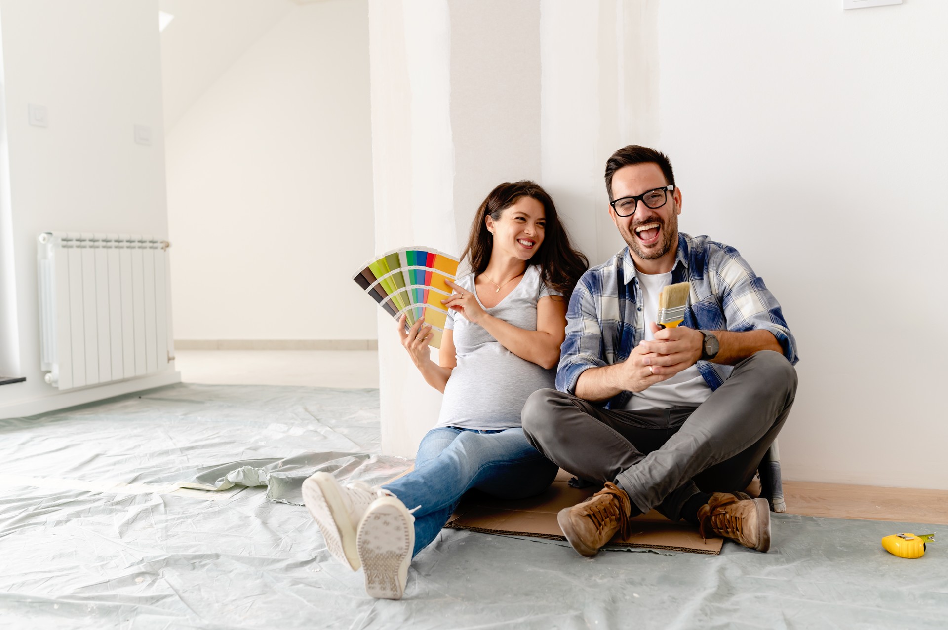 A young, beautiful, and cheerful couple, consisting of a man and his pregnant wife, are seated on the floor of an empty house. They are in the process of selecting colors for decorating their new home